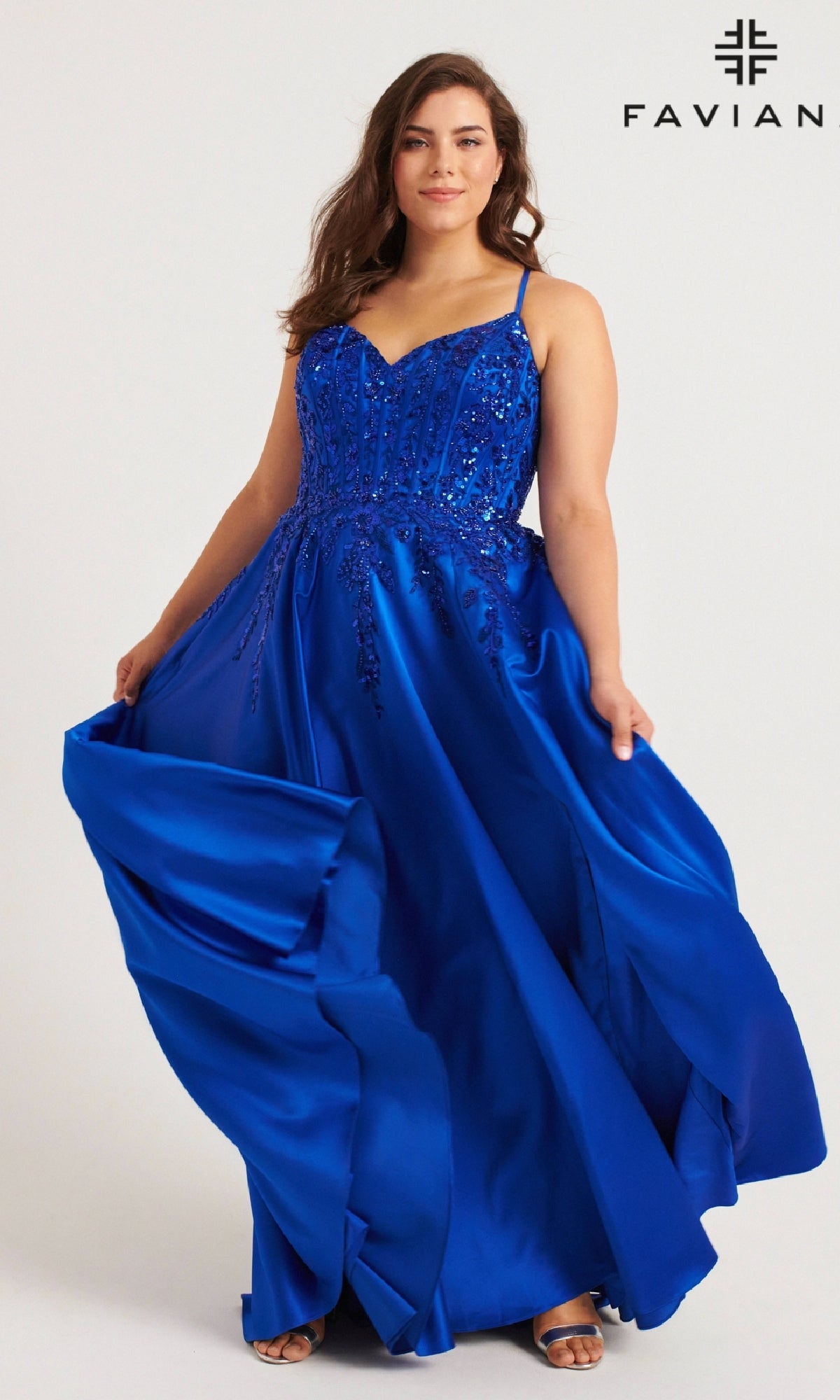 A woman poses against a light background wearing the Faviana 9575 Long Plus-Size Prom Dress—a vibrant blue, beaded corset gown with flowing fabric—with the Faviana logo in the corner.